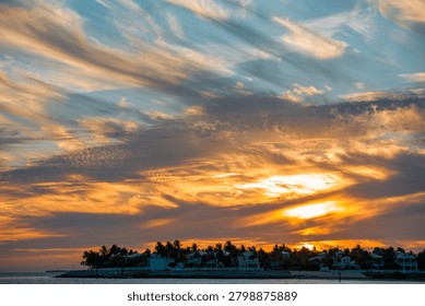 The wide cirrus cloud formations completely cover the colorful evening sky over the dark silhouette of Key West during the summer sunset. The dark silhouettes of the resort houses and the palm trees stand on the Florida ocean coast against the glowing sky.