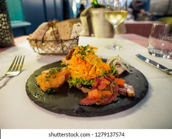 Wide Closeup Of A Fancy Entree Of Smoked Salmon, Foie Gras, And Salad Served On A Slate Plate At A Gourmet Restaurant With Fine Wine. Colmar, France. Travel And Gourmet Dining.