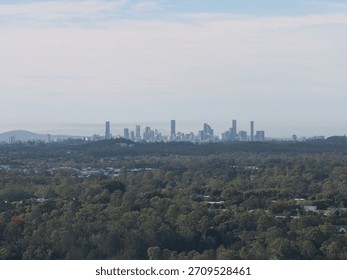Wide elevated view of Brisbane with a dense green forested area and suburban housing in the foreground and the city skyline in the distance under a partly cloudy sky.