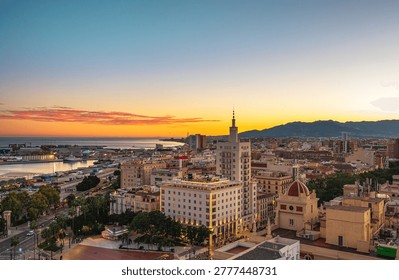 Wide elevated view of Malaga, Spain showing the city skyline, port and waterfront at sunset, with mountains silhouetted under a clear colorful sky. Streets, squares, palm trees and varied urban buildings are visible in the foreground.