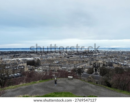 Wide elevated view over Edinburgh showing historic buildings, distant Firth of Forth and two visitors on a hilltop.