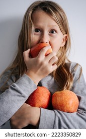 Wide Eyed Boy Biting On An Apple, Holding A Bunch In His Arm. Over White Wall. He Has Long Blond Hair.