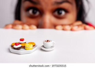 Wide Eyed Hungry Young Woman Staring At A Plate Of Cookies And Mug Of Coffee With Yearning In A Fun Image Using Miniature Toy Food And Cappuccino With Focus To The Refreshments