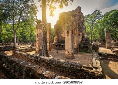 A wide front view capturing the large ruined brick mandapa structure, showcasing the remaining ancient sitting Buddha statue on the left and the prominent standing Buddha statue in the central alcove at Wat Phra Si Iriyabot (Temple of the Four Postures) in the UNESCO-listed Kamphaeng Phet Historical Park, Thailand.