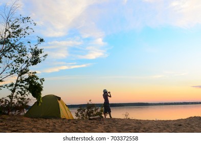 Wide Girl In Yoga Pants And A Hat Outdoors On A Sandy Beach By The River And Tourist Tents By The Tree And Bushes Taking Pictures On The Phone Beautiful Sunset Sky With Clouds.