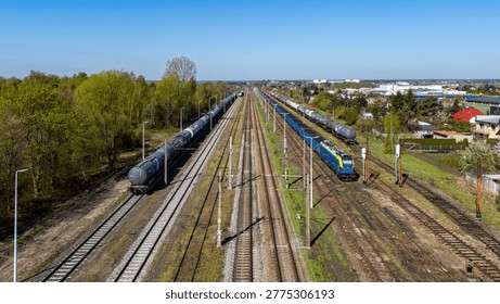 Wide high-angle aerial view of a multi-track railway with freight trains, blue and yellow electric locomotive, tank wagons and cargo cars, surrounded by green trees and suburban houses under clear blu