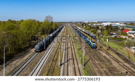 Wide high-angle aerial view of a multi-track railway with freight trains, blue and yellow electric locomotive, tank wagons and cargo cars, surrounded by green trees and suburban houses under clear blu