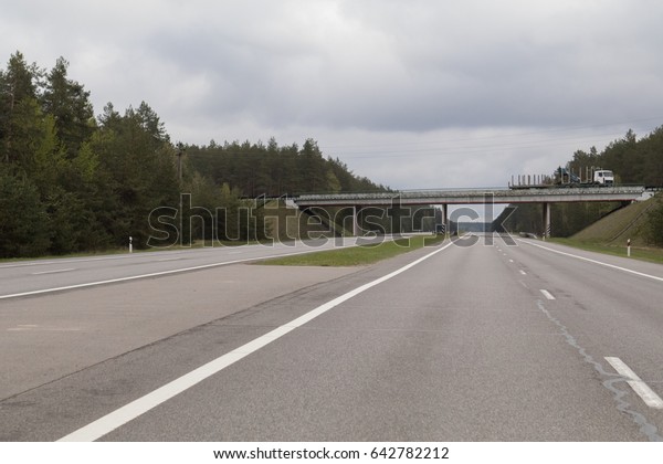 A wide highway, a bridge, green trees and a dark\
sky with gray clouds