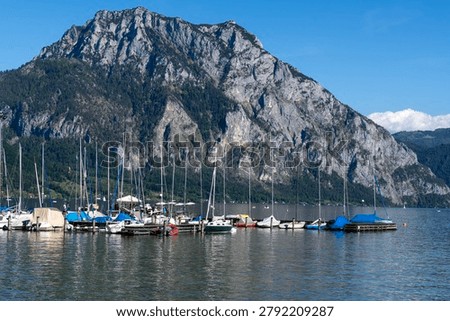 A wide horizontal landscape capturing a bustling lakeside harbor filled with many modern yachts and sailboats at a dock in Austria. The foreground highlights the calm water surface reflecting the boats, while the breathtaking background features a towering mountain massif with massive grey cliffs and patches of green forest under a clear blue summer sky. Ideal for regional travel promotion, luxury lifestyle advertising, and alpine nautical sport concepts.
