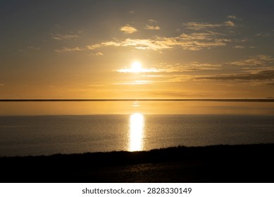 Wide horizontal landscape shot of a dramatic golden sunset reflecting brightly over the calm ocean surface and coastal waters along the southern shore of Iceland.