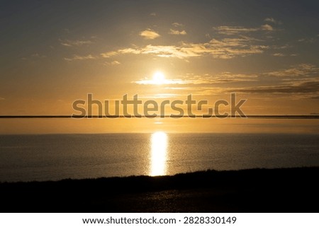 Wide horizontal landscape shot of a dramatic golden sunset reflecting brightly over the calm ocean surface and coastal waters along the southern shore of Iceland.