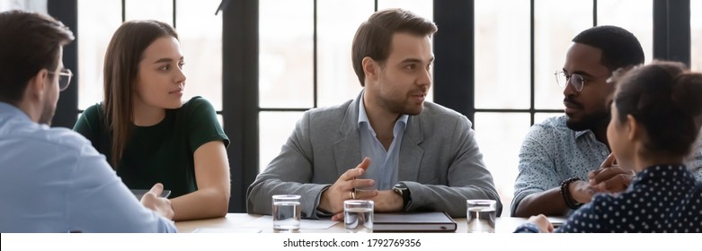 Wide Image Confident Businessman Speaking, Sharing Opinion, Discussing Project At Corporate Meeting With Diverse Colleagues, Sitting At Table In Boardroom, Leader Business Coach Training Staff