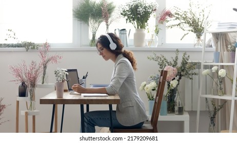 Wide image of young Caucasian female florist sit at desk in atelier wear headphones study online on computer. Woman decorator work in earphones in own studio, consult client customer on web on laptop. - Powered by Shutterstock