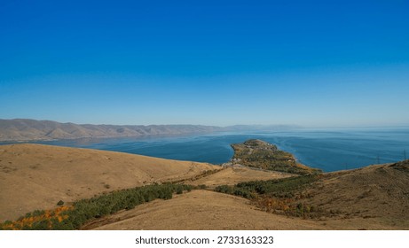 Wide landscape of Lake Sevan, Armenia. The calm lake stretches beneath clear blue skies, surrounded by dry hills and mountain ridges in the distance.