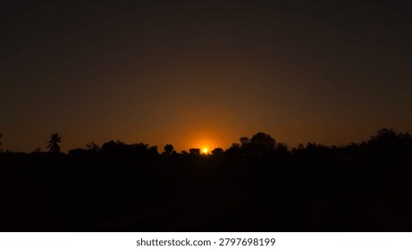 A wide landscape shot capturing a dramatic sunset over a dark silhouette of trees and distant fields. The bright sun is dipping just below the horizon, emitting a brilliant golden-orange glow that softly fades into a vast, dark, clear evening sky. The foreground and the line of trees are completely cast in shadow, emphasizing the peaceful transition from day to night in a rural or natural setting