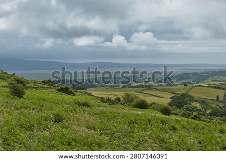 A wide landscape view of rolling green hills and patchwork fields leading to an estuary. Distant Cumbrian mountains rise under a cloudy sky.