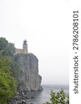 Wide landscape view of Split Rock Lighthouse above the waters of Lake Superior under an overcast sky and light mist. Historic landmark integrated into a natural coastal scene with the lighthouse as the visual focal point.