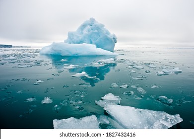 A wide low angle view of melting sea ice floes in still waters of Northern Arctic with iceberg and glacial wall in background.Climate Crisis and Breakdown.Climate emergency