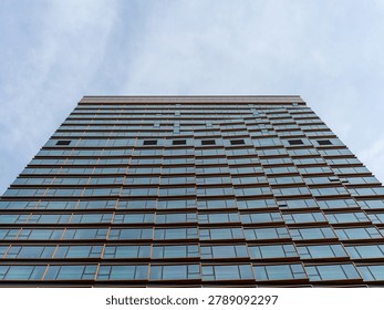 Wide low-angle perspective looking straight up at a massive, symmetrical modern glass skyscraper against a cloudy sky. The composition represents urban density, corporate power, architectural engineering, and financial success.