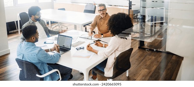 In a wide office shot, four professionals are deep in discussion around a table, showcasing a dynamic and collaborative team in action. A diverse team in a friendly and inclusive workplace atmosphere - Powered by Shutterstock