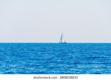 Wide open sea view with a tiny white sailboat on a hazy horizon under a pale blue sky, minimal seascape, summer sailing, Mediterranean travel.