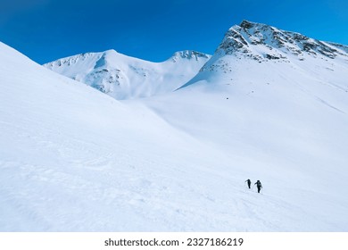 Wide open shot of two skiers walk up on randoneering skis on steep snowy mountain slope. Extreme sport adventure, ski touring experience on sunny mountain face. Riding fresh powder snow