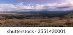 Wide panorama of Chuya steppe in the twilight evening: hilly landscape, endless fields with yellow grass, golden autumn in Altai. In the distance is a mountain ridge with snow covered mountain peaks