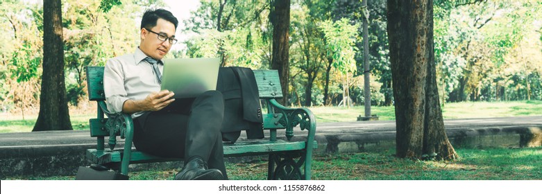 Wide Panorama Confident Asian Businessman Sitting On Bench Working With Laptop Computer At Natural Park, Handsome Young Man Relaxing After Work, Panoramic Banner Asia Business People Working Outside
