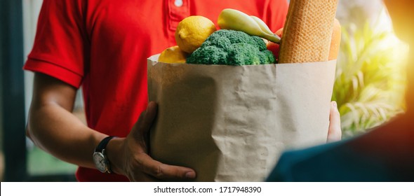 Wide Panoramic Banner Of Woman Customer Receiving Fresh Food Set Bag From Food Delivery Service Man With Protection Face Mask In Red Uniform At Door Home, Express Delivery And Food Delivery Concept