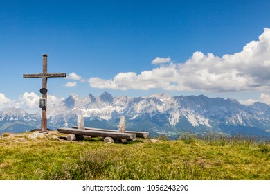 Wide Panoramic Landscape View From Summit Of Mountain Gasselhoehe With Cross And Wooden Bench To Mountain Range Dachstein In Styria, Austria