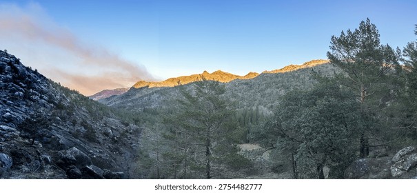 A wide panoramic view captures the first light hitting rugged mountain ridges. Soft morning glow contrasts with deep shadows over the rocky slopes and evergreen pine trees in the Geres valley.