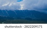 A wide panoramic view of a cloud-capped mountain ridge with visible snow patches, overlooking a dense pine forest below, a small green road sign faintly visible at the tree line