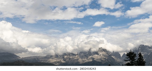 Wide panoramic view of dramatic cloud formations rolling over the rugged Dolomite mountain range in northern Italy. Scenic alpine landscape with snow-dusted peaks, dynamic weather and expansive blue sky in the Italian Alps.