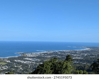 A wide panoramic view from an elevated escarpment lookout showing the full sweep of Wollongong's coastal suburbs nestled between the ocean and the Illawarra Escarpment. The curved coastline with breaking surf, tree-lined suburbs, and the deep blue Pacific Ocean stretch to the horizon under a clear blue sky.