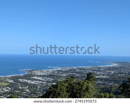 A wide panoramic view from an elevated escarpment lookout showing the full sweep of Wollongong's coastal suburbs nestled between the ocean and the Illawarra Escarpment. The curved coastline with breaking surf, tree-lined suburbs, and the deep blue Pacific Ocean stretch to the horizon under a clear blue sky.