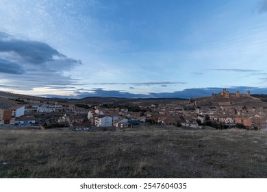 Wide panoramic view of a historic Spanish village with a hilltop castle and expansive countryside under a dramatic evening sky. - Powered by Shutterstock