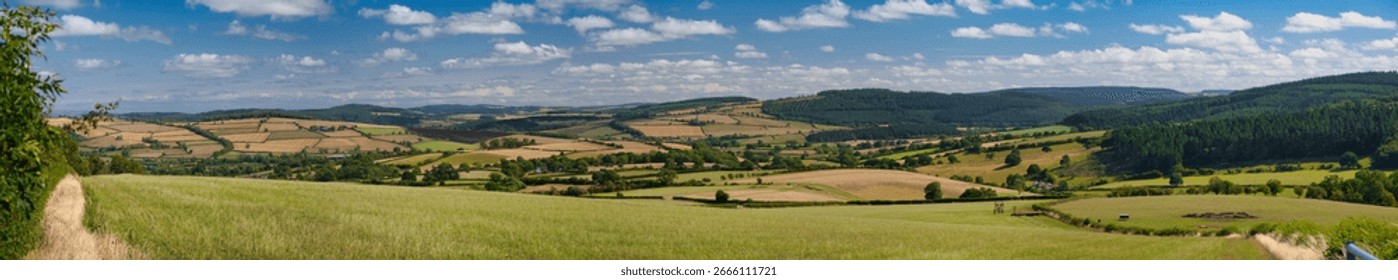 Wide panoramic view of patchwork fields and rolling hills under blue skies in the Shropshire Hills National Landscape, Shropshire, UK. Bright summer light and fluffy clouds convey a fresh rural feel.