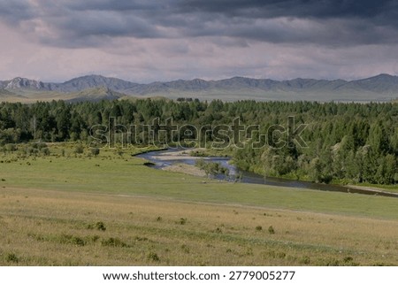 Wide panoramic view of a winding river, green meadow and dense forest with distant mountains under dramatic storm clouds in the Altai region, Russia