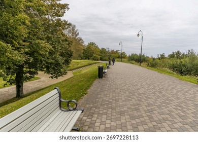 A wide paved park path lined with modern street lamps and empty benches, with a lone person walking in the distance under an overcast autumn sky.