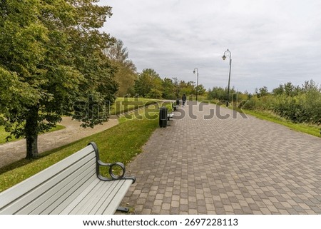 A wide paved park path lined with modern street lamps and empty benches, with a lone person walking in the distance under an overcast autumn sky.