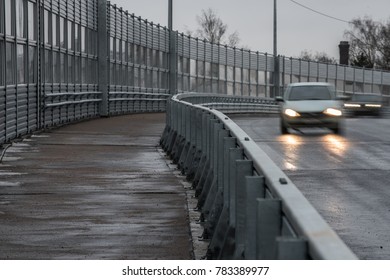 A wide pedestrian road is left along the high-speed viaduct. Silhouettes of two cars on the road. Powerful anti-noise protective fences along the highway.