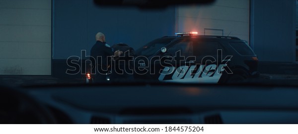 WIDE Police officer searches and handcuffs a\
suspect, African-American Black criminal. Police car lights\
flashing in the background