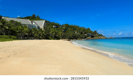 Wide pristine sandy beach next to clear turquoise sea, with modern luxury resort buildings nestled in lush green tropical hillside under clear blue sky