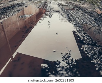 A wide, reflective puddle dominates a muddy, tire-tracked rural road, offering a clear mirror image of distant structures under a soft sky. Perfect for concepts of duality, journey, or environmental i