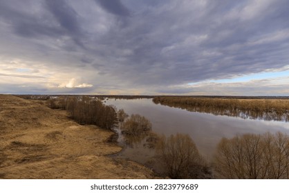 Wide river flows calmly through marshy terrain under dramatic cloudy sky