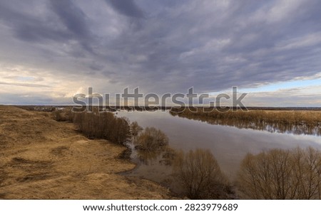 Wide river flows calmly through marshy terrain under dramatic cloudy sky