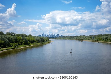 A wide river flows toward a distant urban skyline under a bright, cloud-filled summer sky, framed by lush green banks on both sides. On the captured landscape, the Vistula River stretches horizontally, its calm surface reflecting the deep azure heavens above. Verdant trees densely populate the shorelines, creating a vibrant contrast against the dark water and the sandy edges of the riverbanks. In the background, modern glass skyscrapers and slender high-rises emerge from the horizon, marking the central business district of a major European city. A small, solitary marker buoy floats quietly in the mid-ground, while a collection of specialized technical equipment and orange buoys anchored near the right bank suggests ongoing water management or maintenance activities. The perspective is elevated, likely from a bridge, offering a panoramic view that emphasizes the peaceful harmony between nature and metropolitan development. Fluffy, white cumulus clouds drift lazily across the expanse, casting subtle, shifting shadows over the terrain. This scenery perfectly encapsulates the serene atmosphere of a warm afternoon where urban growth meets a tranquil, natural waterway, inviting quiet contemplation of the peaceful environment.