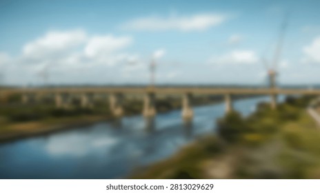 A wide river flows under a long bridge with multiple arches, surrounded by lush green trees and vegetation, under a bright sky with scattered clouds.
