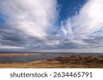 A wide river winds through a flat, open landscape under a dramatic sky filled with scattered clouds.