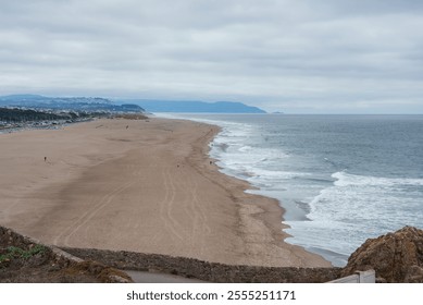 Wide sandy Ocean Beach stretches along the Pacific Ocean with a distant cityscape and hills. Overcast sky and gentle waves create a serene atmosphere. - Powered by Shutterstock